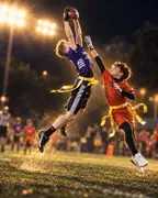 A receiver in purple leaps to catch a pass over a defender in orange, with a referee and families watching